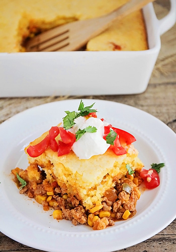 a square of tamale pie made with cornbread and cheese and topped with tomatoes and sour cream on a white plate. A casserole pan with filled with tamale pie that has a square cut out with a wooden spoon in it's place.