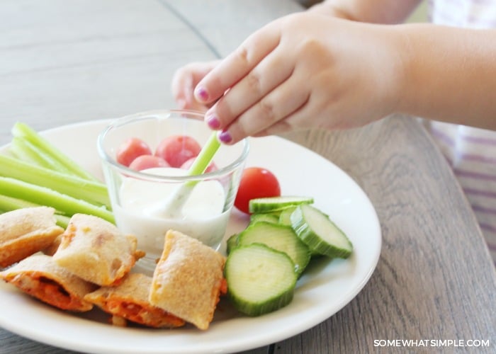 a child's hand dipping a celery stick into a dish of ranch on a plate with other vegetables and pizza bites