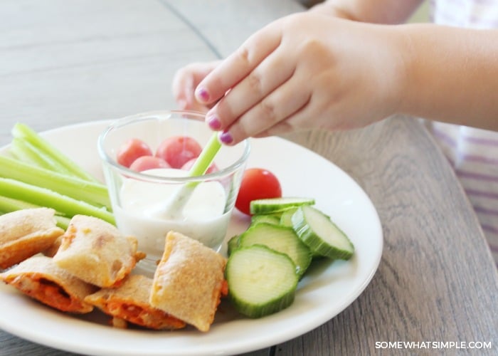 a child's hand dipping a celery stick into a dish of ranch on a plate with other vegetables and pizza bites