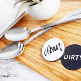 two dishwasher magnets sitting on a cutting board next to two spoons. One side is black and says dirty, the other one is white and says clean