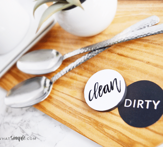 two dishwasher magnets sitting on a cutting board next to two spoons. One side is black and says dirty, the other one is white and says clean