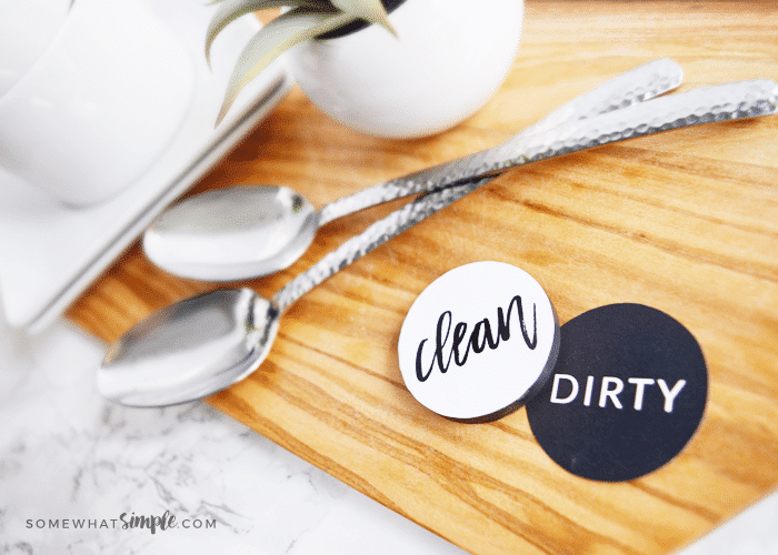 two dishwasher magnets sitting on a cutting board next to two spoons. One side is black and says dirty, the other one is white and says clean