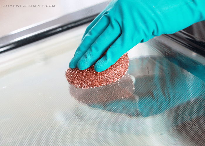 a gloved hand scrubbing a glass oven door with a steel wool sponge is how to clean your oven easily