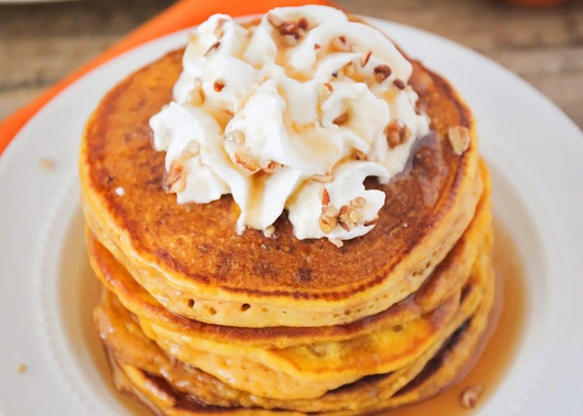 Stack of 3 orange pumpkin pancakes made using the best Pumpkin Pancake Recipe you will find. The pancakes are topped with syrup, whipped cream and nuts on a white plate.