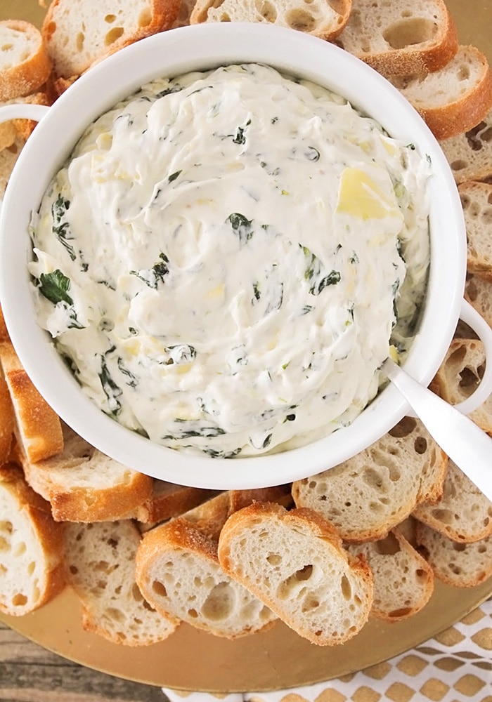 looking down on a white bowl of dip made with spinach and artichoke with slices of bread around the bowl.