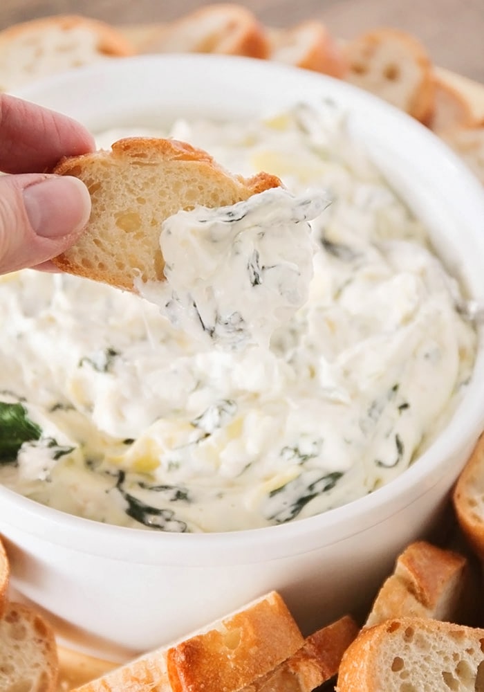 a hand dipping a piece of bread into a hot bowl of spinach artichoke dip