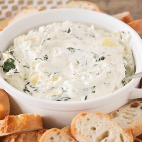 a white bowl filled with Cheesy Spinach Artichoke Dip that has a spoon stuck in the dip. Surrounding the bowl are slices of baguettes.