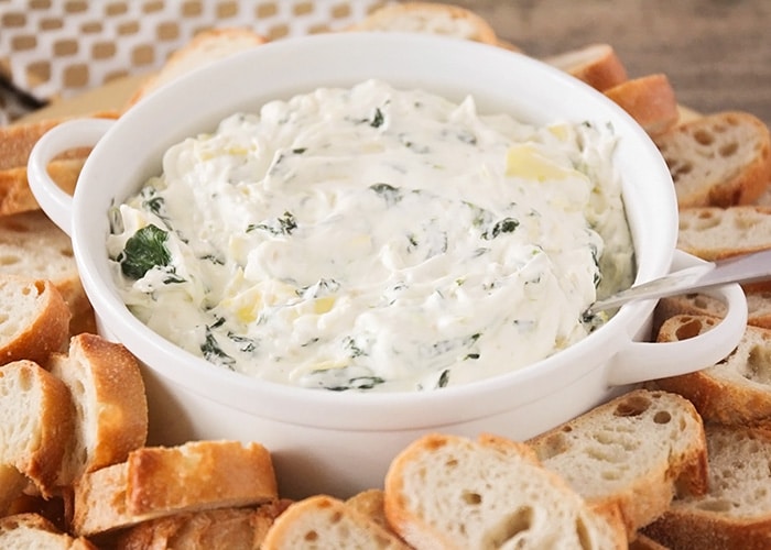 a white bowl filled with Cheesy Spinach Artichoke Dip that has a spoon stuck in the dip. Surrounding the bowl are slices of baguettes.