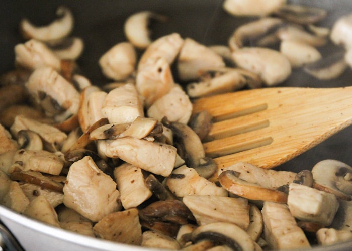 a wooden spoon stirring pieces of chicken and mushrooms in a skillet