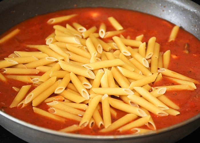 a skillet full of red pasta sauce and with uncooked penne pasta noodles poured into the pan