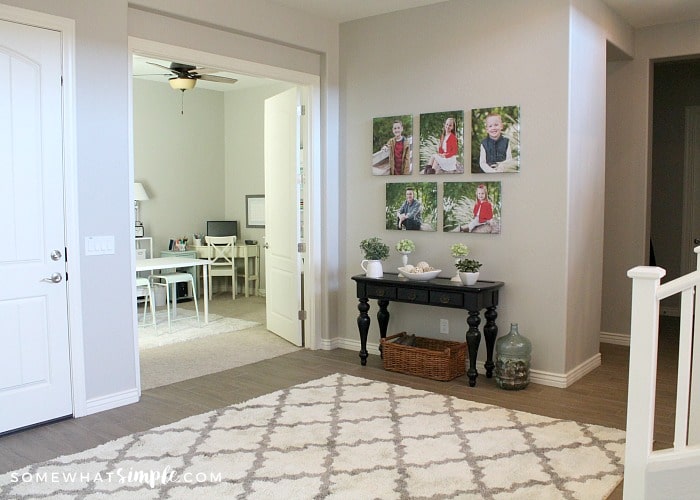 the entry way to a home with a white rug on the floor. There is a small black table with family pictures above it. From this angle you are looking through the entry way into a home office.