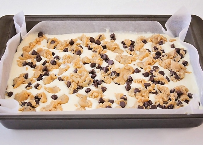 the cookie bar dough base spread out in a parchment paper lined baking pan with a layer of cream cheese over the top