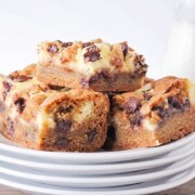 a pile of cheesecake cookie bars on a stack of white plates. Next to the plates are a glass pitcher of mile and a red and white striped straw laying down.