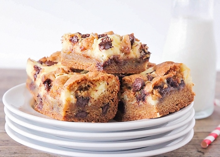 a pile of cheesecake cookie bars on a stack of white plates. Next to the plates are a glass pitcher of mile and a red and white striped straw laying down.