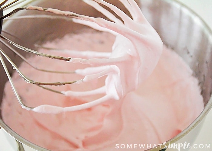 a stand mixer whisk covered in the mixture to make Strawberry Marshmallows over a bowl filled with the pink covered mixture.