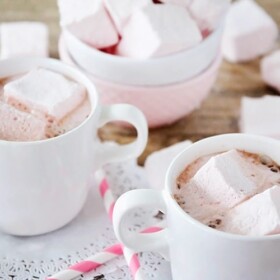 two cups of hot chocolate that have Homemade Strawberry Marshmallows floating in them. Behind the mugs is a bowl of more marshmallows.