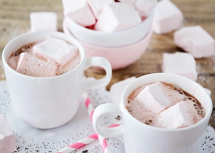 two cups of hot chocolate that have Homemade Strawberry Marshmallows floating in them. Behind the mugs is a bowl of more marshmallows.