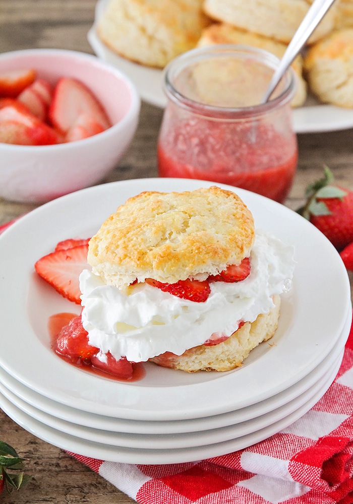 a piece of easy Strawberry Shortcake made using this simple recipe on a white plate. Behind the plate is a plate full of biscuits, a bowl of sliced strawberries and a jar of strawberry sauce.