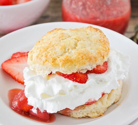 a piece of strawberry shortcake made with a biscuit and filled with sliced strawberries and whipped cream on a white plate. Additional strawberry slices are on the plate.