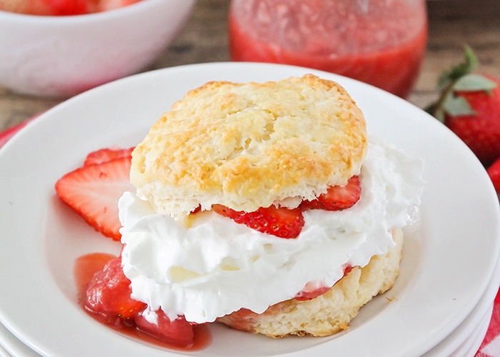 a piece of strawberry shortcake made with a biscuit and filled with sliced strawberries and whipped cream on a white plate. Additional strawberry slices are on the plate.