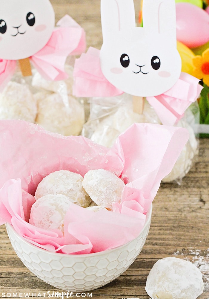 A white bowl filled with pink tissue paper and bunny tail cookies. Behind the bowl are two Easter bunny gifts filled with these easy cookies