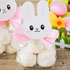 two packages of Easter bunny tail cookies wrapped in plastic with a pink bow and topped with an Easter bunny printable. Next to the cookie gift bags is an Easter basket filled with plastic eggs.