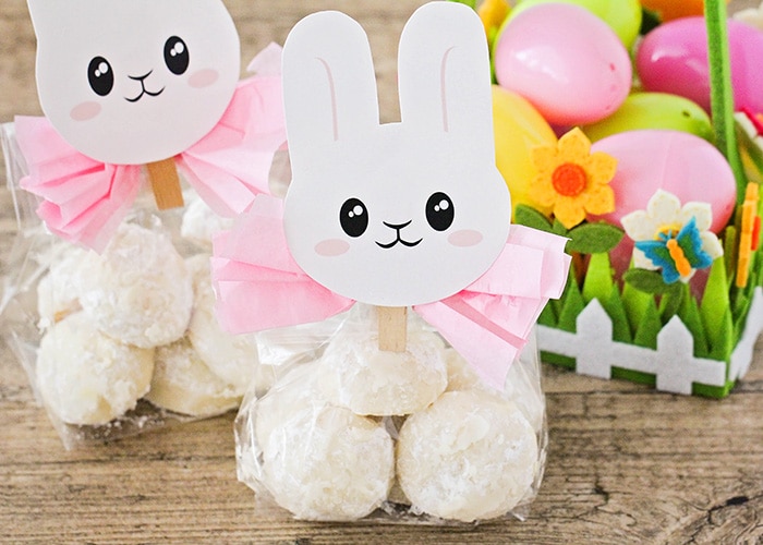 two packages of Easter bunny tail cookies wrapped in plastic with a pink bow and topped with an Easter bunny printable. Next to the cookie gift bags is an Easter basket filled with plastic eggs.