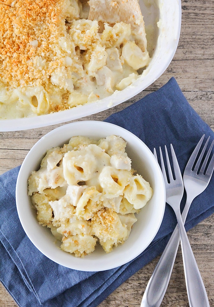 looking down on a white bowl filled with baked tortellini casserole that is topped with bread crumbs. The bowl is sitting on a blue cloth napkin and two forks are next to the bowl. Behind the bowl is a casserole filled with this tortellini recipe.