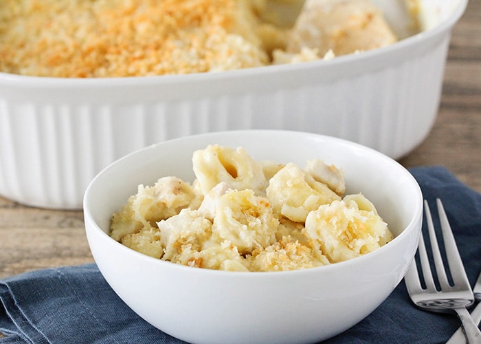 a white bowl filled with cheese baked tortellini topped with bread crumbs. The bowl is sitting on a blue cloth napkin and a fork is next to it. Behind the bowl is a casserole filled with this tortellini recipe.