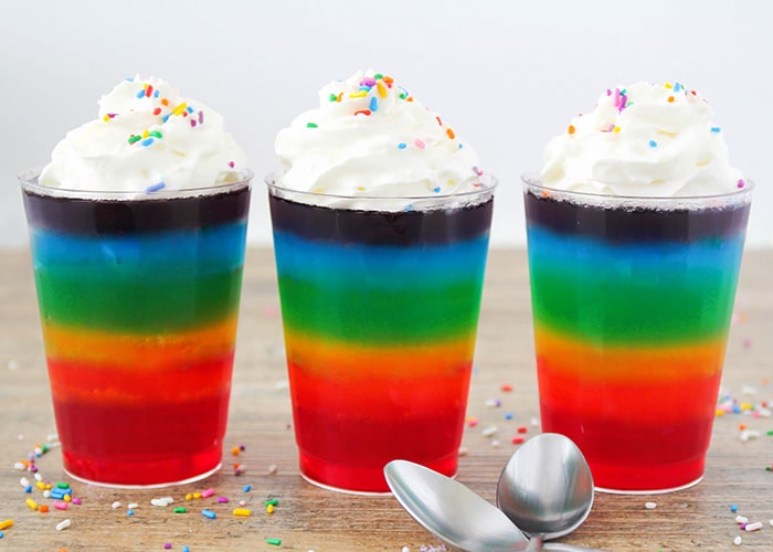 three cups with layered rainbow jello topped with whipped cream and sprinkles. Starting from the top, the jello layers are purple, blue, green, yellow, orange and red.