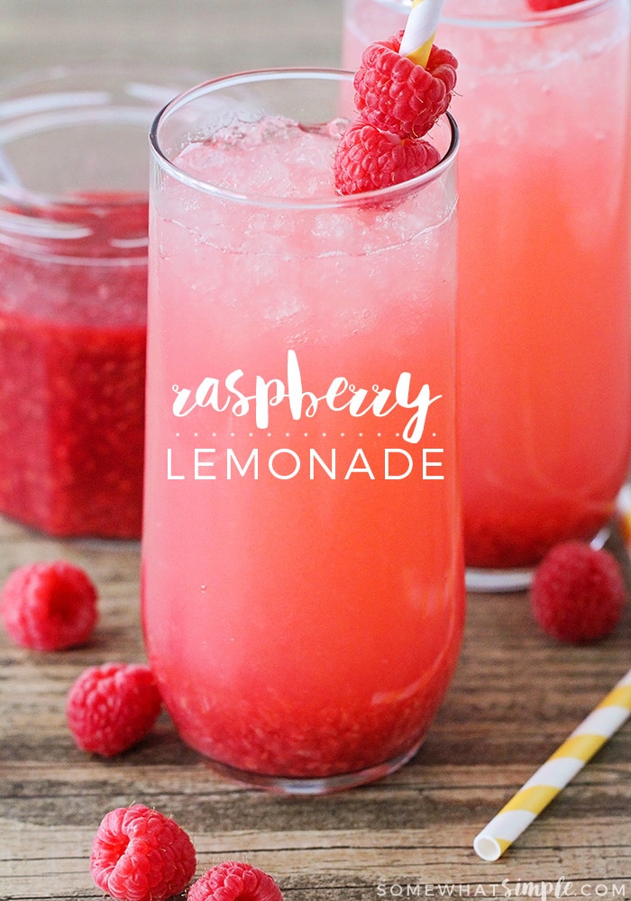 a glass of raspberry lemonade made with this simple recipe. Another glass of lemonade, a jar of raspberry sauce are in the background and three raspberries and a colored straw are lying on the table.