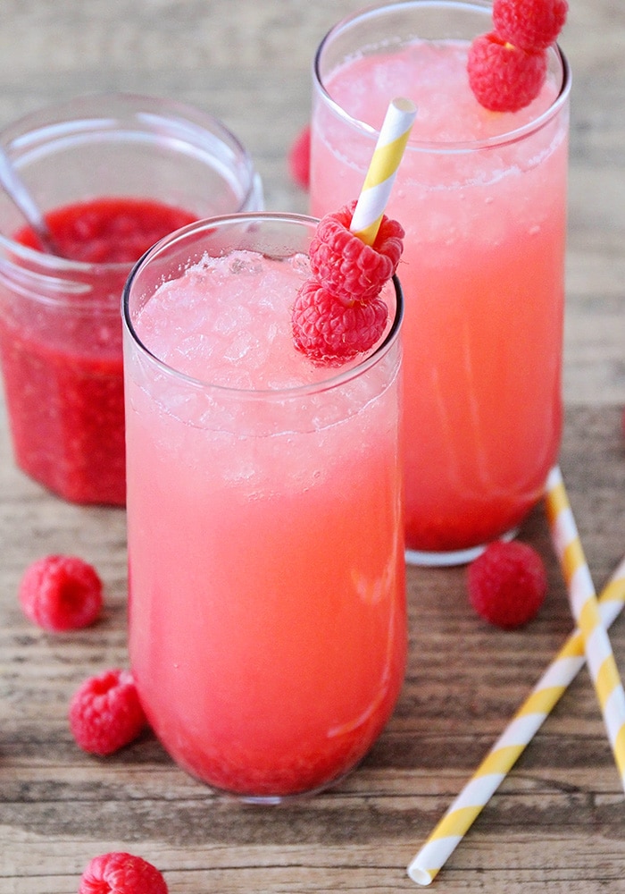 two glasses filled with raspberry lemonade with yellow striped straws and raspberries wrapped around the straws. A jar of raspberry sauce is on the table next to the glasses.