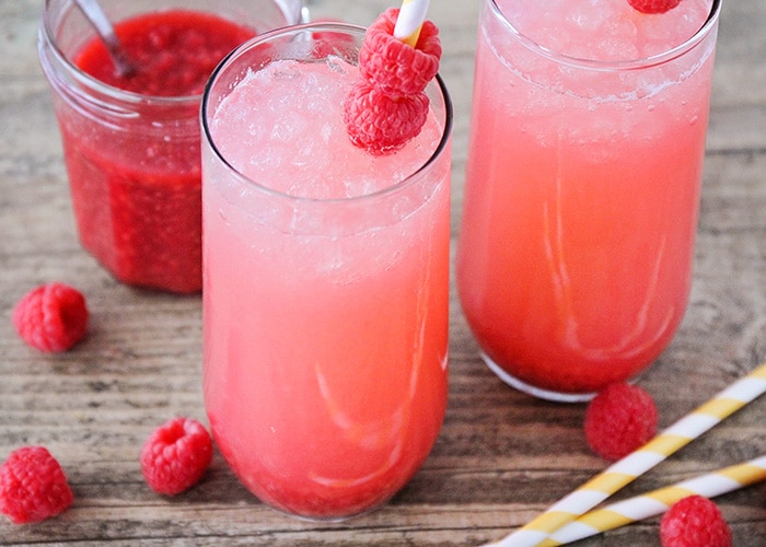 two tall glasses filled with raspberry lemonade and each has a straw that has been slide through two raspberries 