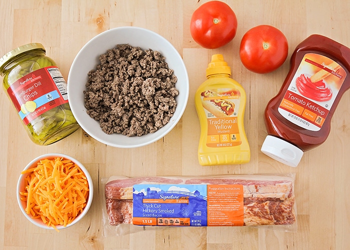 looking down on the ingredients needed to make this bacon cheeseburger pizza recipe; ground beef, pickles, cheese, bacon mustard, ketchup and tomatoes