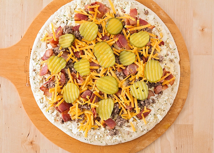 looking down on a raw pizza crust with cheese ground beef, bacon cheese and pickles on top before it is placed in the oven