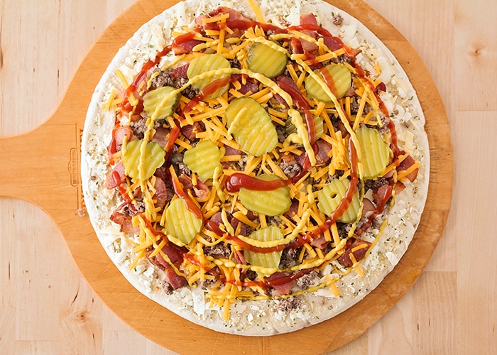 looking down on a raw pizza crust with cheese ground beef, bacon cheese and pickles with a swirl of ketchup and mustard on top before it is placed in the oven
