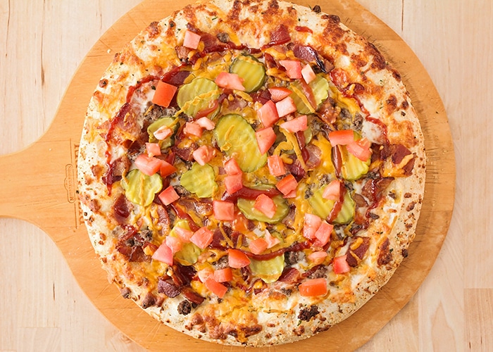 looking down on a baked bacon cheeseburger pizza with cheese ground beef, bacon cheese and pickles with a swirl of ketchup and mustard on top before it is placed in the oven