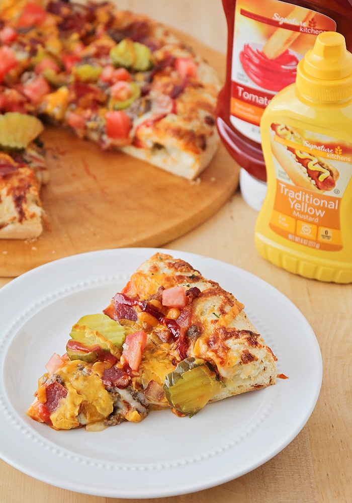 a slice of bacon cheeseburger pizza on a white plate with the rest of the cheeseburger pizza and a bottle of ketchup and mustard in the background