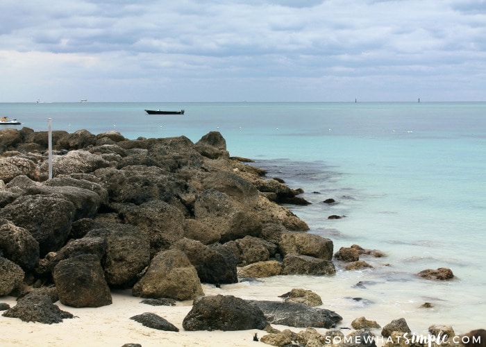 a view of a white sand beach in Freeport, Bahamas that has large rocks at the end of the beach. There are few small boats in the water off in the distance.