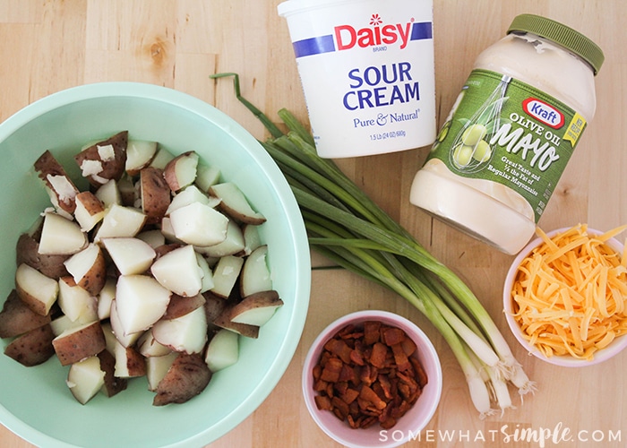 a bowl of cut potatoes in a blue bowl, a jar of mayonnaise, a container of sour cream, a bowl of chopped bacon, a bowl of cheddar cheese and a few green onion stalks on a table which are the ingredients to make a loaded baked potato salad