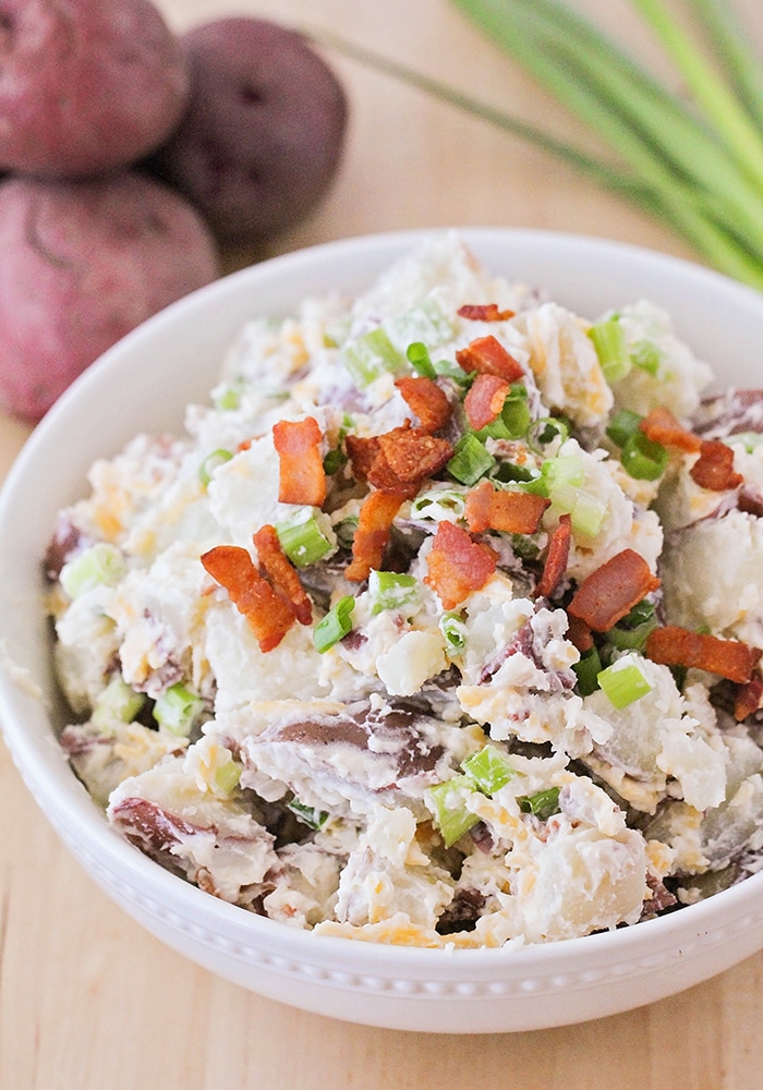 a close up of a white bowl filled with this easy baked potato salad recipe topped with cheese, bacon and green onions with three red potatoes and stalks of green onions in on the table in the background