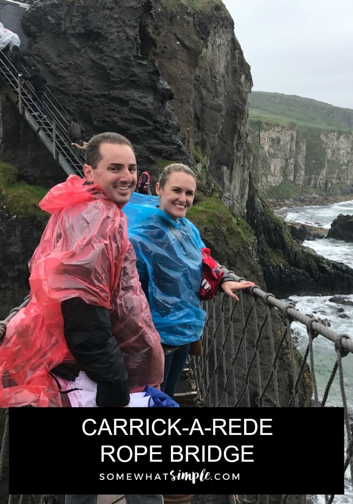 a good looking couple wearing rain ponchos standing on the carrick-a-rede suspension bridge with the ocean and cliffs in the background