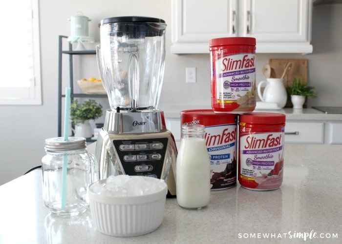 a blender sitting on a counter next to a mason jar glass, a jar of milk, a bowl of ice and cans of chocolate, strawberry and vanilla slim fast shake powders.