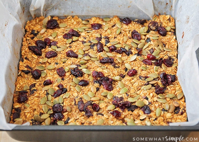 a square baking pan line with parchment paper and filled with ingredients including granola, dried cranberries and pumpkin seeds that has just been baked in the oven