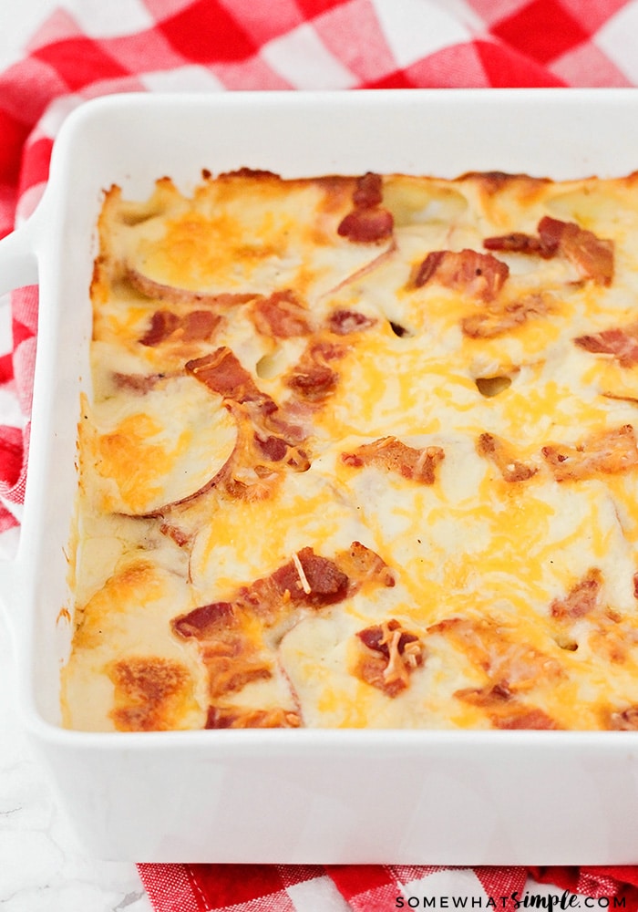 a full white backing pan of scalloped potatoes that are fresh out of the oven and are a golden brown and topped with pieces of bacon and cheddar cheese. a red checkered cloth napkin is laying on the counter above the pan.