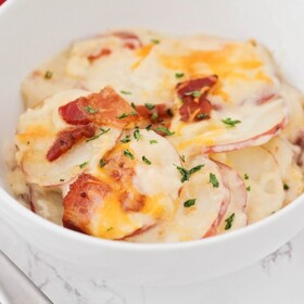 looking down on a white bowl filled about halfway with scalloped potatoes and topped with cheddar cheese, bacon and parsley. A fork is next to the bowl and a red checkered cloth napkin is laying behind the bowl.
