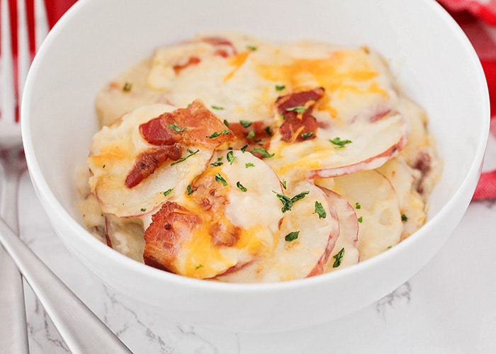 looking down on a white bowl filled about halfway with scalloped potatoes and topped with cheddar cheese, bacon and parsley. A fork is next to the bowl and a red checkered cloth napkin is laying behind the bowl.