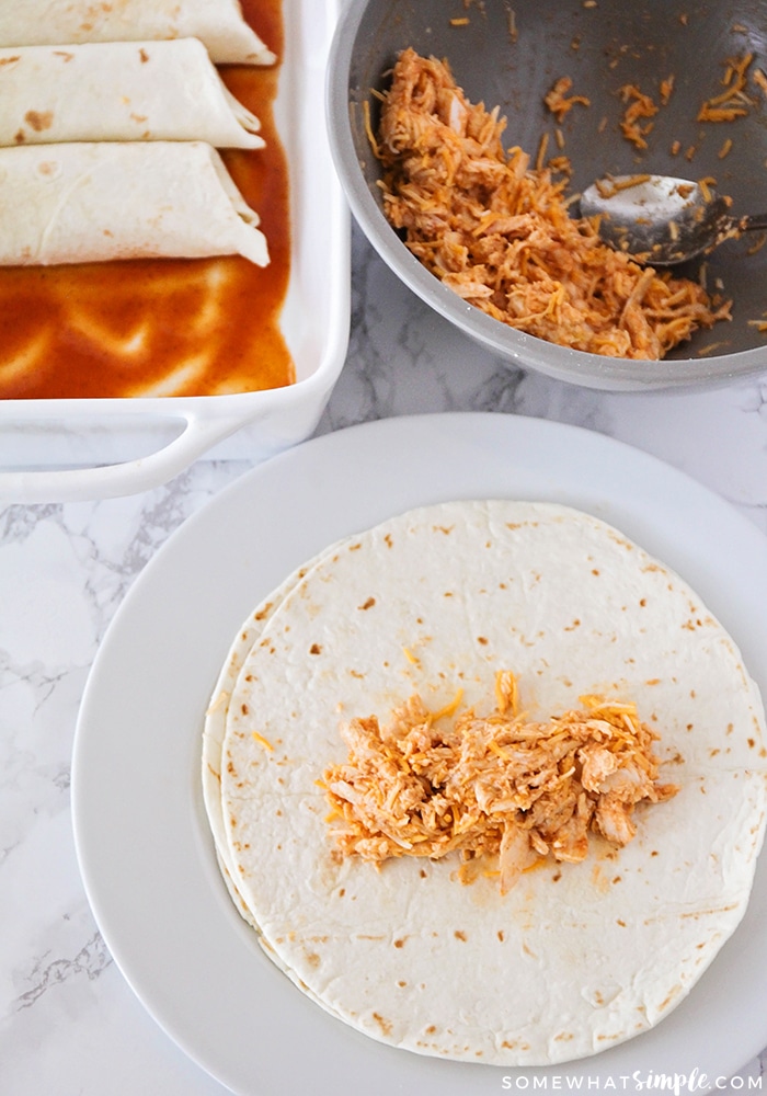 a tortilla with chicken on it and a bowl with the chicken mix in the back and a tray filled with a few chicken enchiladas