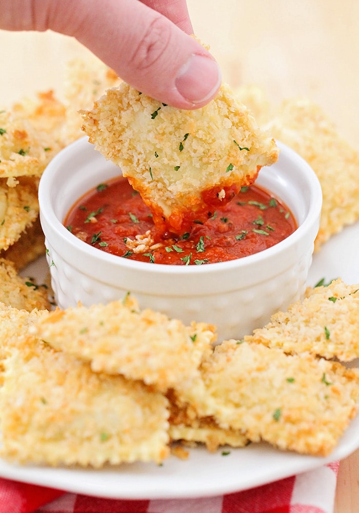 a toasted ravioli being dipped in marinara sauce with several toasted raviolis on a plate around the dipping sauce