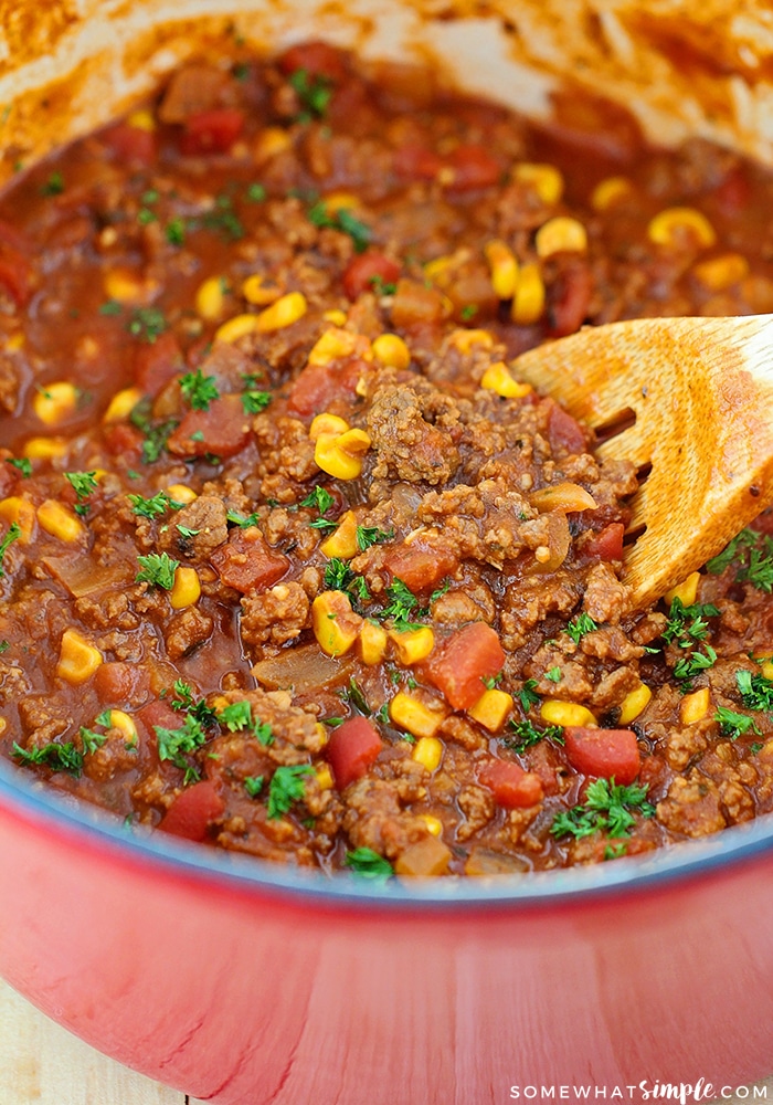 a red pot full of this easy Goulash recipe with a wooden spoon stirring the ground beef, corn, tomatoes and other ingredients